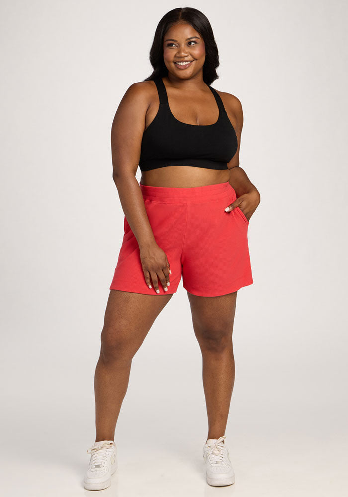 Smiling woman in a black sports bra, Woolx Maisey Ribbed Shorts - Hibiscus, and white sneakers stands hand-in-pocket. | Sapphire is 5’8”, wearing a size XL