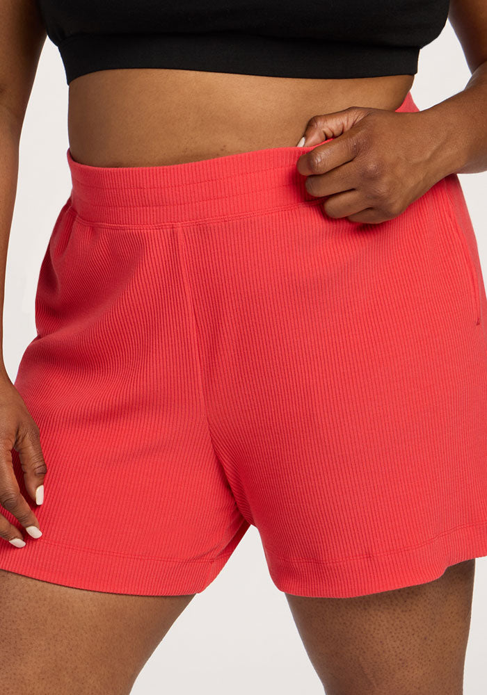 Person wears Woolx Maisey Ribbed Shorts in Hibiscus with a black crop top, holding the waistband.