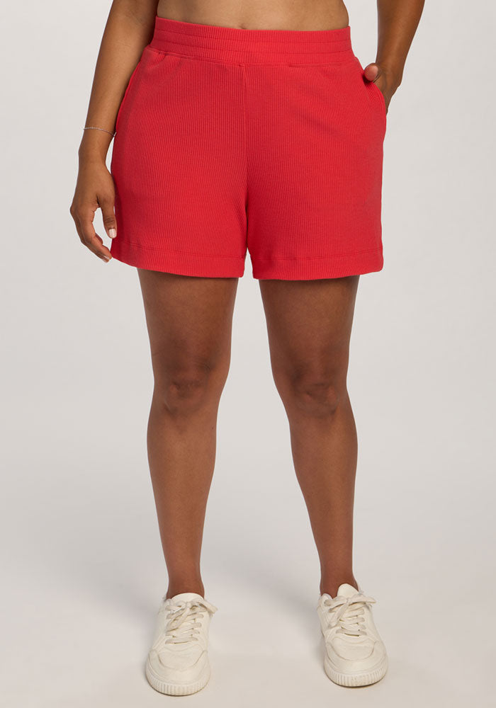 A person in Woolx Maisey Ribbed Shorts - Hibiscus, a tan top, and white sneakers stands with hands in pockets.
