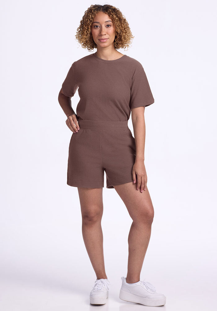 Smiling woman with curly hair wears Woolx Maisey Ribbed Shorts in Peppercorn and a brown tee against a white background.