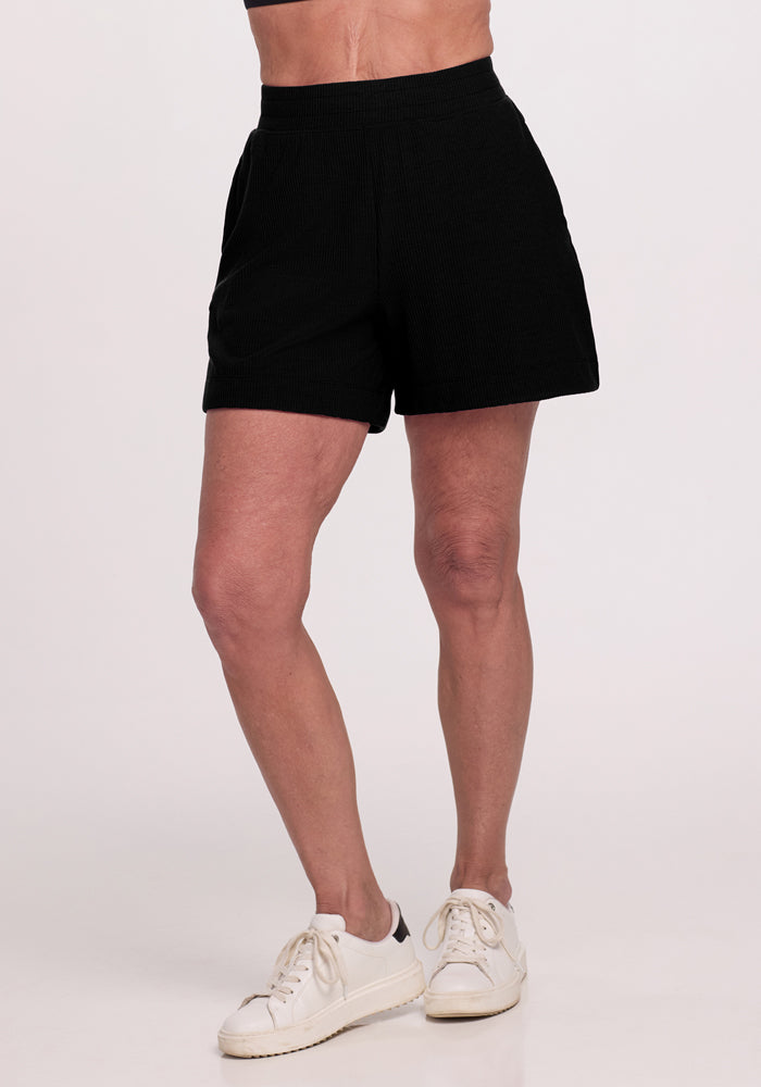 A person is wearing Maisey Ribbed Shorts in black by Woolx and white sneakers, standing with legs slightly apart against a plain white background, with their hands resting at their sides. | Tam is 5’8.5”, wearing a size S