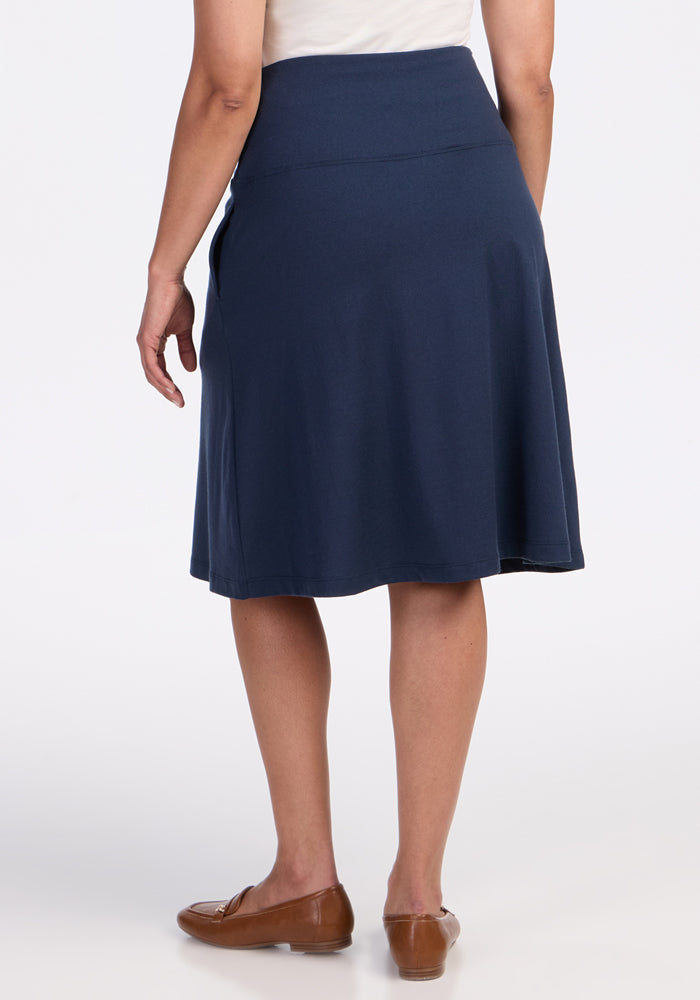 A person is shown from the back against a white background, wearing the Woolx Marilyn Flowy Midi Skirt in Deep Navy, paired with a white top and brown flats; one arm is slightly bent.