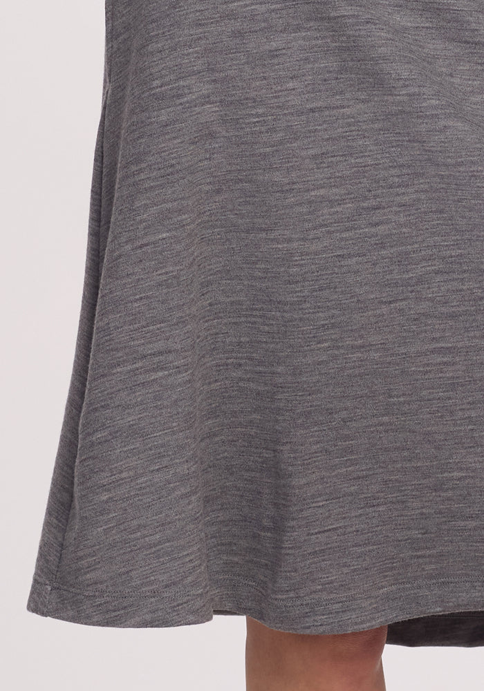 A close-up shows the bottom of the Marilyn Flowy Midi Skirt in Graphite Heather by Woolx, made from textured merino wool. It drapes gently with part of a person's knee visible at the blurred bottom edge against a light, neutral background.