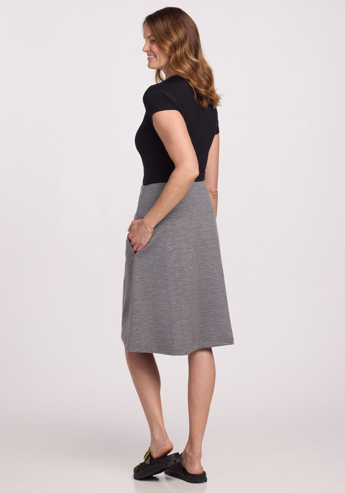 A woman with wavy hair stands in profile, wearing a black top and the Woolx Marilyn Flowy Midi Skirt in Graphite Heather. Her right hand casually rests in her pocket as she slightly smiles, completing her chic look with black shoes against a white background.