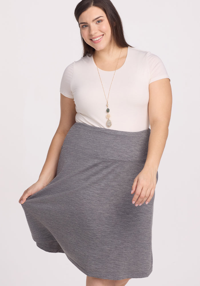 A smiling woman in a white top and long necklace poses while holding the hem of the Marilyn Flowy Midi Skirt by Woolx in Graphite Heather. The A-line silhouette enhances her grace against the plain white background.