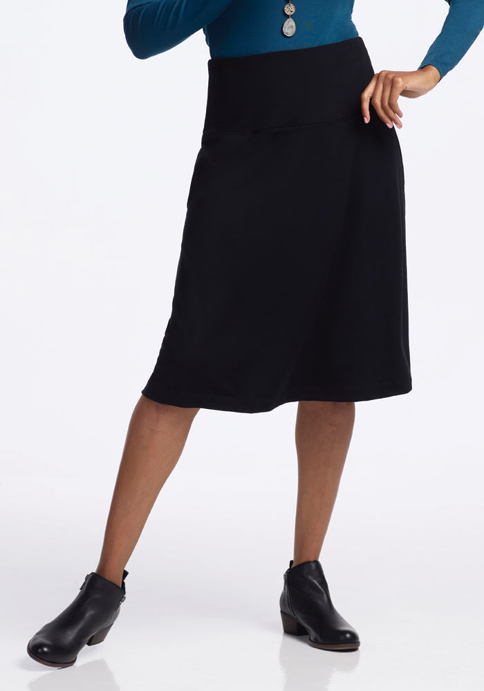 A person wearing a blue long-sleeve top and the Woolx Marilyn Flowy Midi Skirt - Black stands confidently in black ankle boots, one hand on their hip. | Keesha is 5’9.5”, wearing a size S