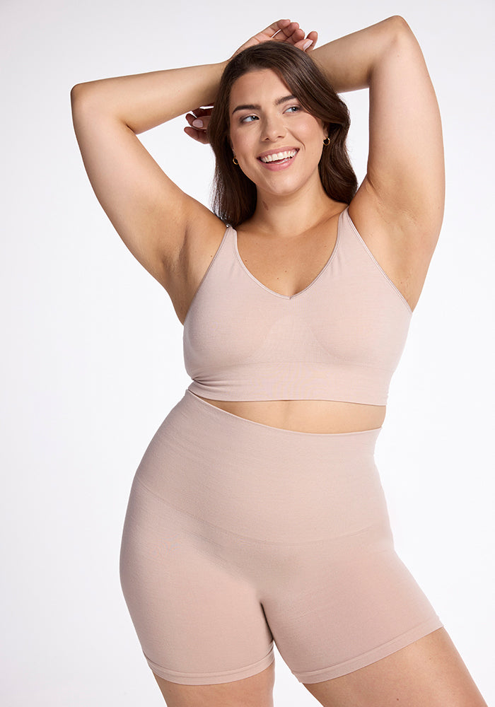 Wearing the Marley Shapewear Shorts in Sand by Woolx, a woman smiles confidently as she highlights the gentle compression of her outfit. One arm is raised while her other hand rests on her head against a plain white background. | Juliana is 5'10", wearing a size XL