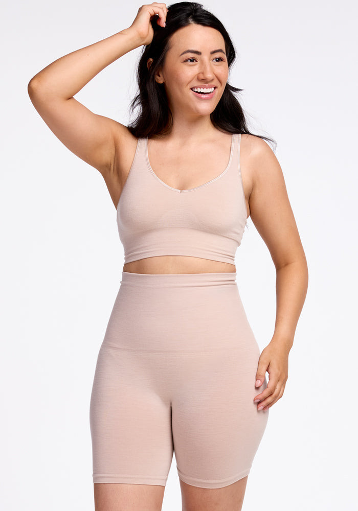 A person with long dark hair, smiling and wearing a light beige tank top along with Marley Shapewear Shorts in Sand by Woolx, stands against a plain white background. The outfit's seamless construction exudes comfort and athletic vibes. | Sarah is 5'8", wearing a size S