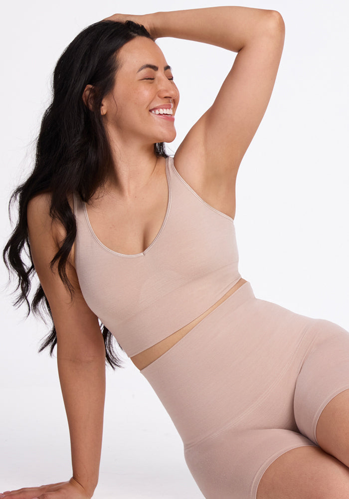 A woman with long dark hair sits on the floor against a plain white background, smiling with her eyes closed. She wears the Woolx Marley Shapewear Shorts in Sand—a high-waisted, seamless set paired with a matching sports bra.