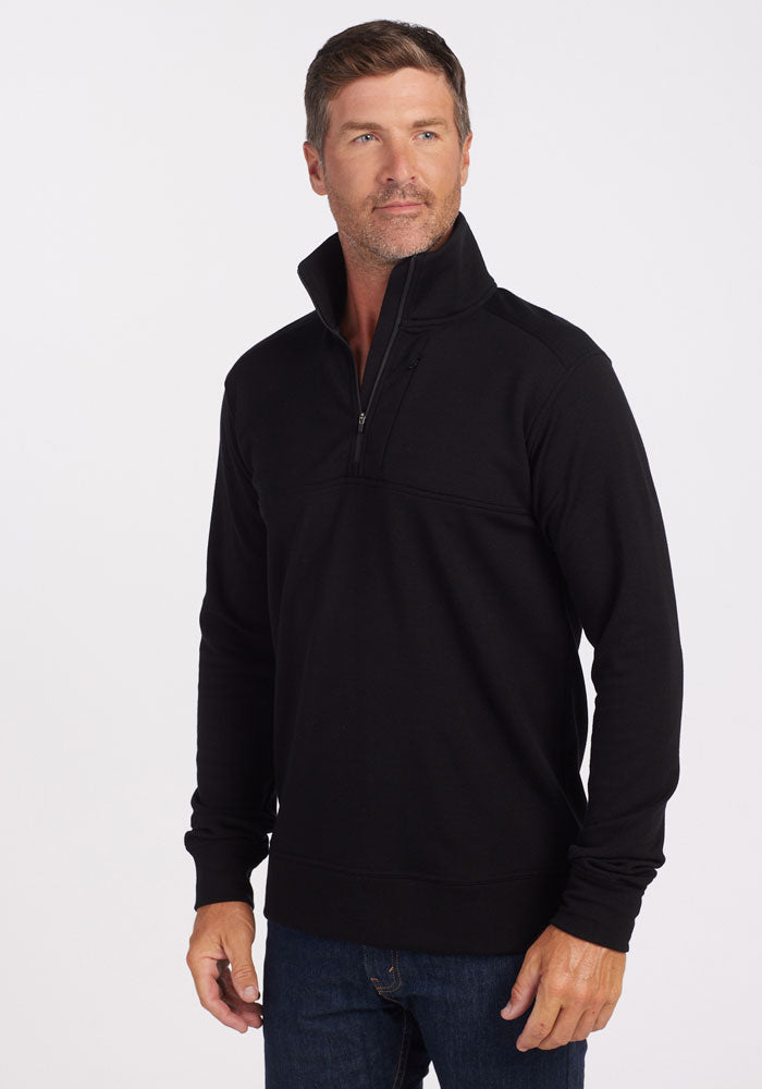 A man wearing a black Woolx Mason 1/4 Zip and blue jeans stands against a white background, looking slightly to his left with a neutral expression. The Merino wool fleece is odor-resistant for all-day freshness.