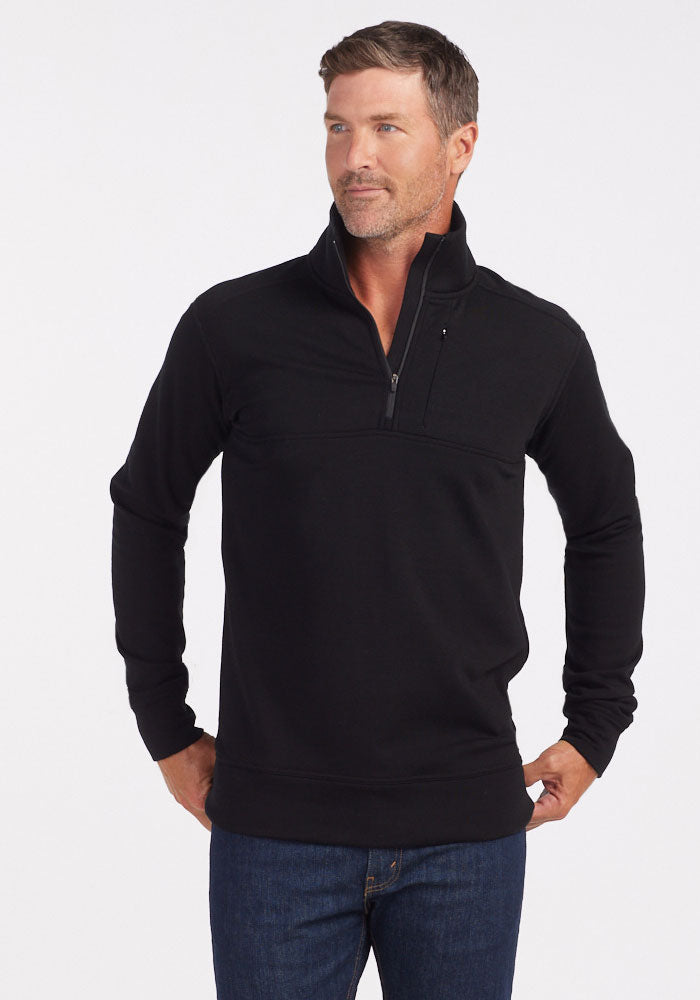 A man in a Woolx Mason 1/4 Zip - Black and blue jeans stands with hands on hips, looking to the side against a white background; the odor-resistant Merino wool fleece provides comfort and style. | Lee is 6’, wearing a size M