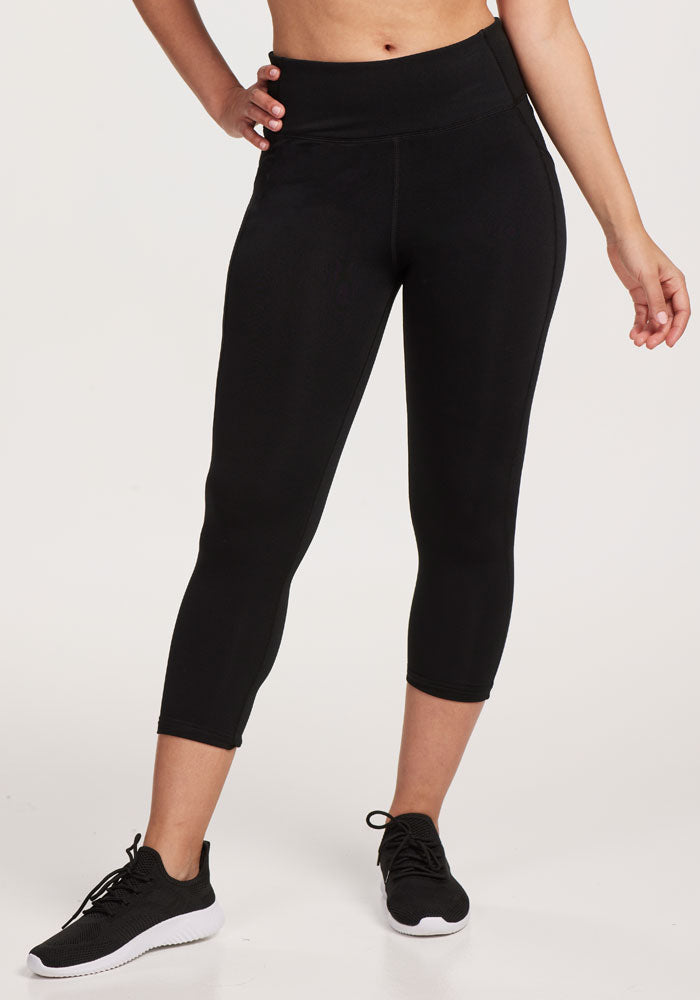 A person models Woolx Merino Flex® McKenna Capris in black, paired with black athletic shoes, standing against a plain white background, one hand on their hip and the other arm relaxed.