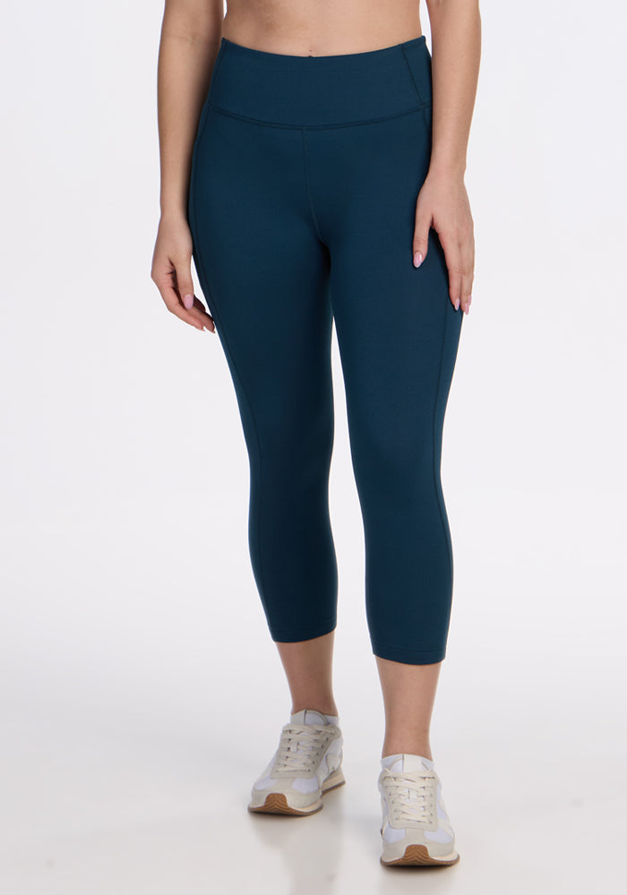 A person models Woolx Merino Flex® McKenna Capris in Majolica Blue, featuring moisture-wicking fabric, paired with white sneakers. Only the lower torso and legs are shown against a plain white background. | Ariana is 5'11", wearing a size S