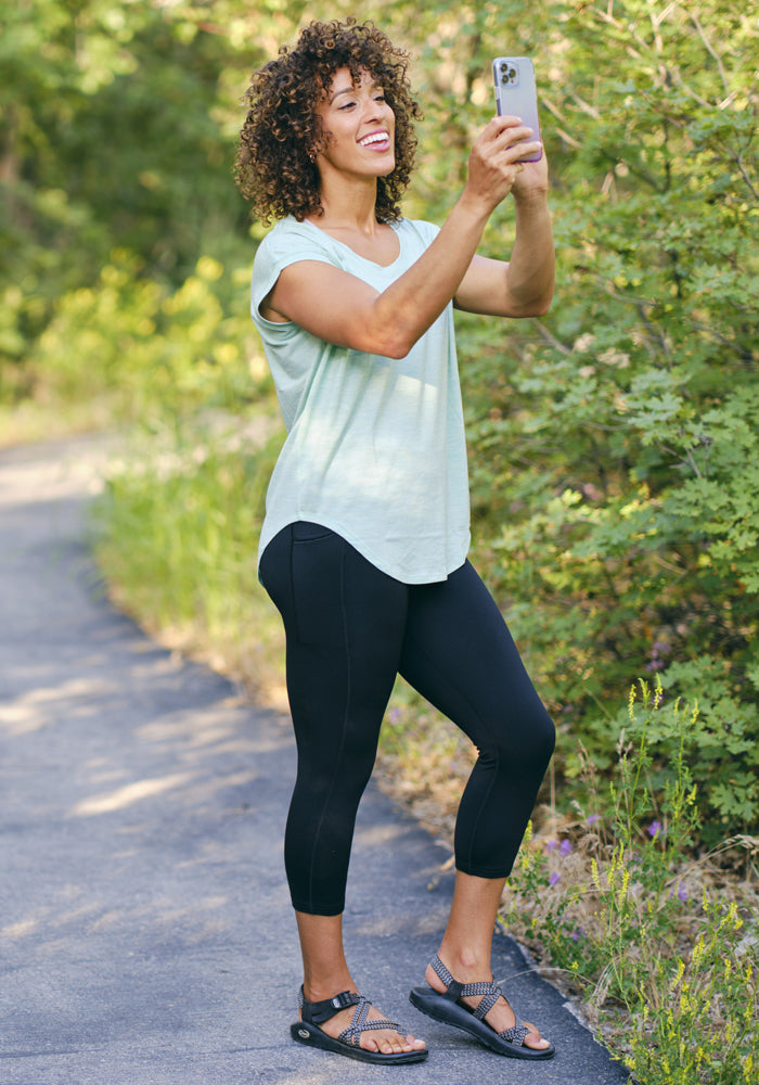 A woman with curly hair smiles for a selfie on a paved path, wearing a light green t-shirt, sandals, and Woolx McKenna Capris—high-waisted leggings made from breathable, moisture-wicking fabric—surrounded by greenery.
