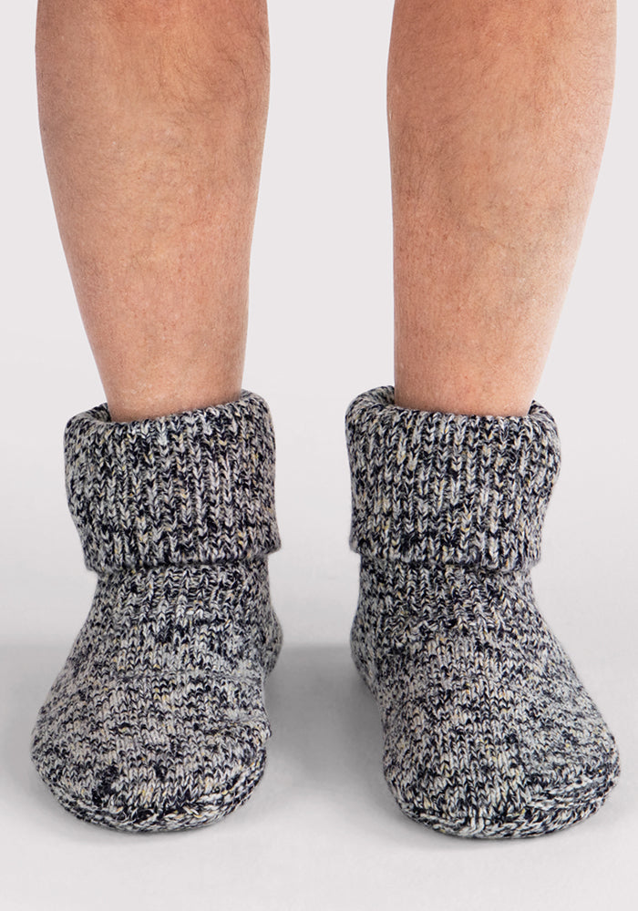 A close-up shows a person wearing Woolx Men's Gripper Slipper Bootie in Speckled Stone, featuring thick gray Merino wool with folded cuffs and plush Sherpa lining, standing on a plain, light-colored surface.