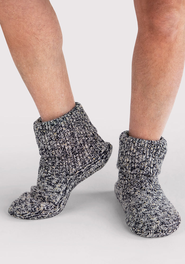 Bare feet wearing Woolx Men's Gripper Slipper Bootie in Speckled Stone, featuring thick marled Merino wool and a fold-over cuff, stand on a light surface.