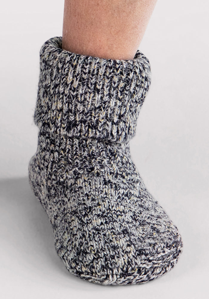 A close-up of a foot wearing the Woolx Men's Gripper Slipper Bootie in Speckled Stone, featuring a cozy, thick design with a folded cuff, against a plain light background.
