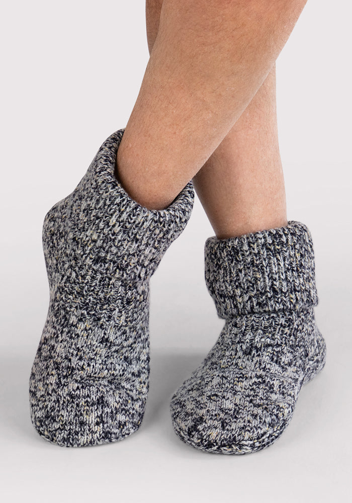 A person wearing Woolx Men's Gripper Slipper Bootie in Speckled Stone stands on a light background with legs crossed at the ankles.