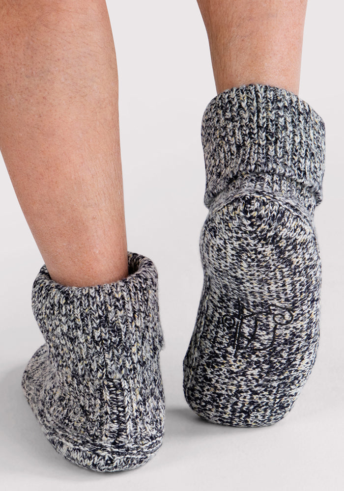 A close-up shows a person wearing Woolx Men's Gripper Slipper Bootie in Speckled Stone, featuring gray-and-white cozy fabric, rubberized grip soles, and plush Sherpa lining, standing on a light background.