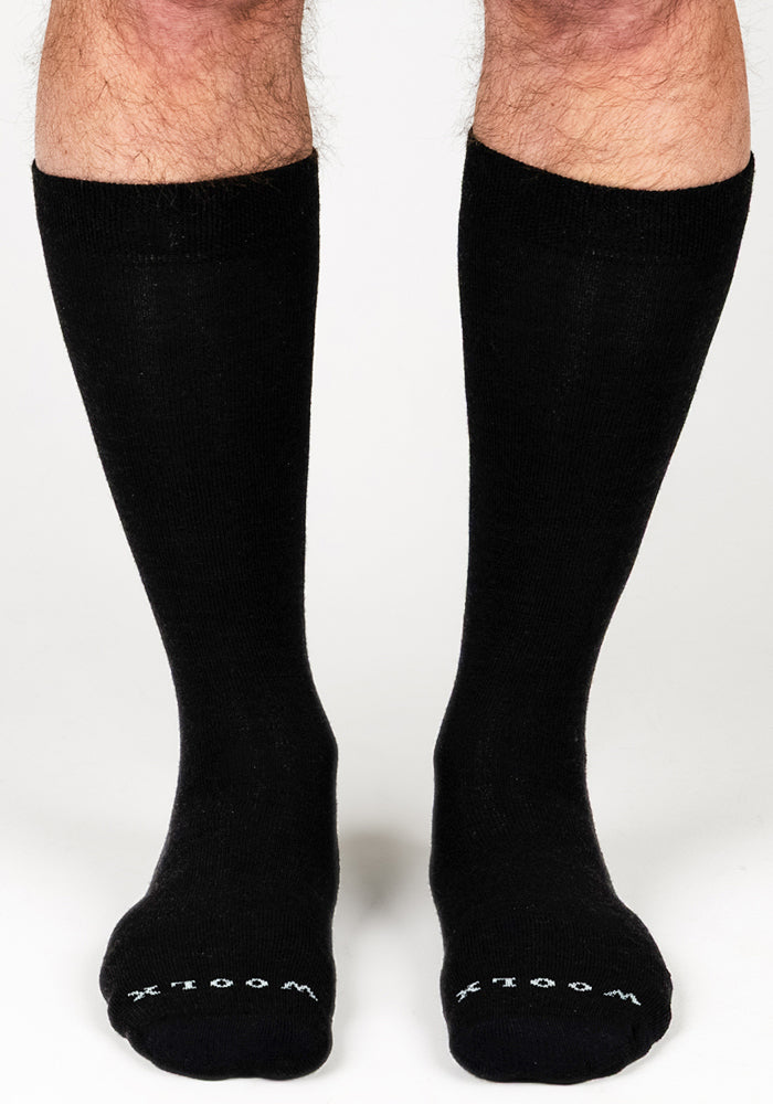 A pair of legs wears Mens Chandler Mid Calf Socks in black by Woolx, featuring "WOOLX" in white on the toes. These Merino wool dress socks are shown against a plain white background.
