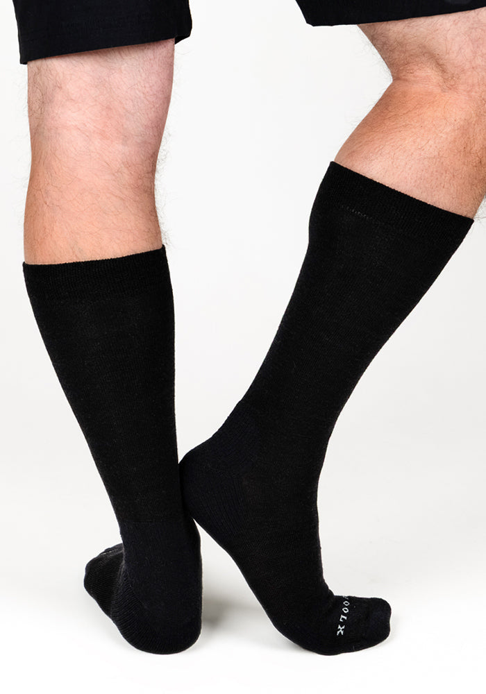 A person models Woolx Mens Chandler Mid Calf Socks in black, standing with legs crossed against a plain white background. Only the legs and part of black shorts are shown.