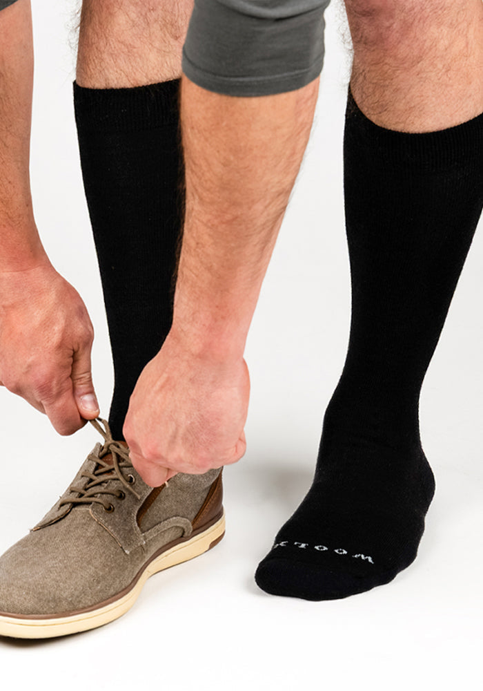 A person wearing Woolx Mens Chandler Mid Calf Socks in black and brown lace-up shoes is tying one shoe, while their other foot, in a sock with "SOCK" partially visible, rests on the ground.