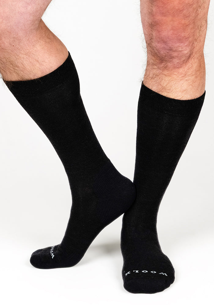 A person wears Woolx Mens Chandler Mid Calf Socks in black, standing with one foot crossed behind the other. The socks have white text on the toes and are displayed against a plain white background.