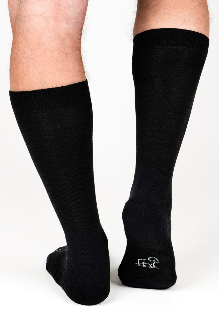 A pair of legs wearing Woolx Mens Chandler Mid Calf Socks in black, shown from the back. One sock features a small white embroidered animal near the heel against a plain white background.