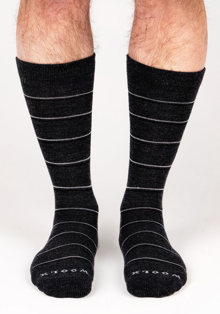 A pair of feet wearing Woolx Mens Chandler Mid Calf Socks in dark grey charcoal, with thin white stripes and "WOOLX" labeled on the toes, are standing on a white surface.