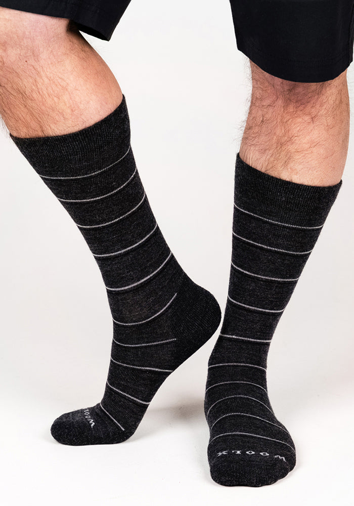 A person stands on a white background, legs slightly bent and dressed in black shorts, wearing Woolx Mens Chandler Mid Calf Socks in dark grey charcoal.