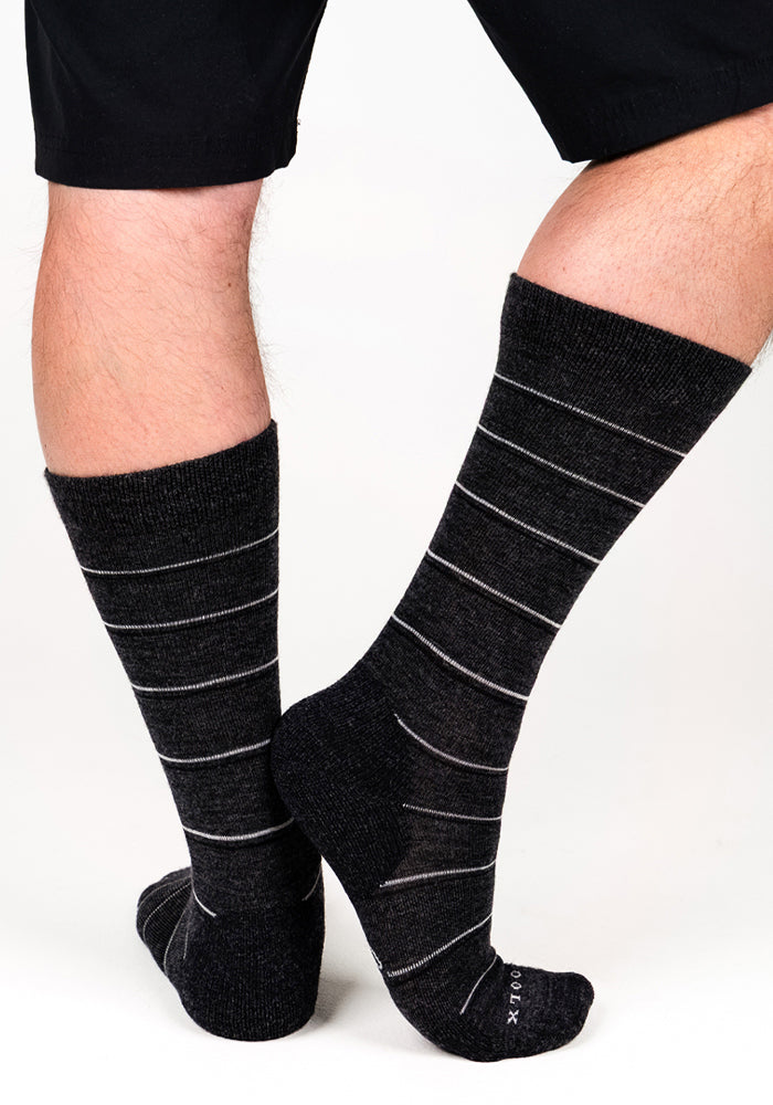 A person wearing black shorts and a pair of Woolx Mens Chandler Mid Calf Socks in Dark Grey Charcoal stands on a white background, with one foot flat and the other raised onto its toes.