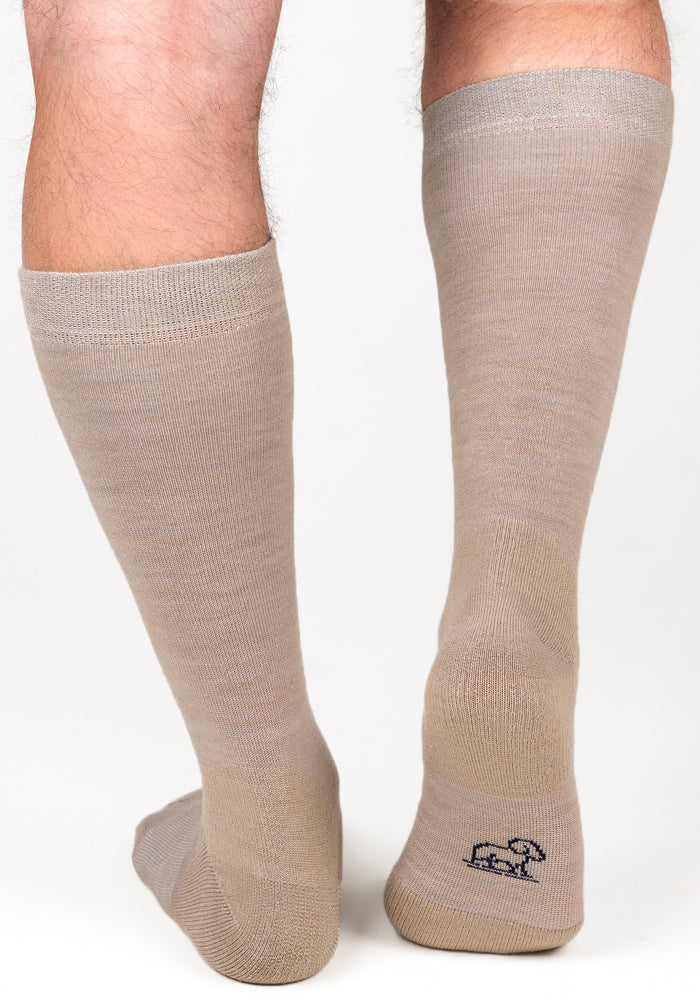A person wears Woolx Mens Chandler Mid Calf Socks in Simply Taupe, standing on a white background. One sock displays a small black outline of an elephant near the heel.