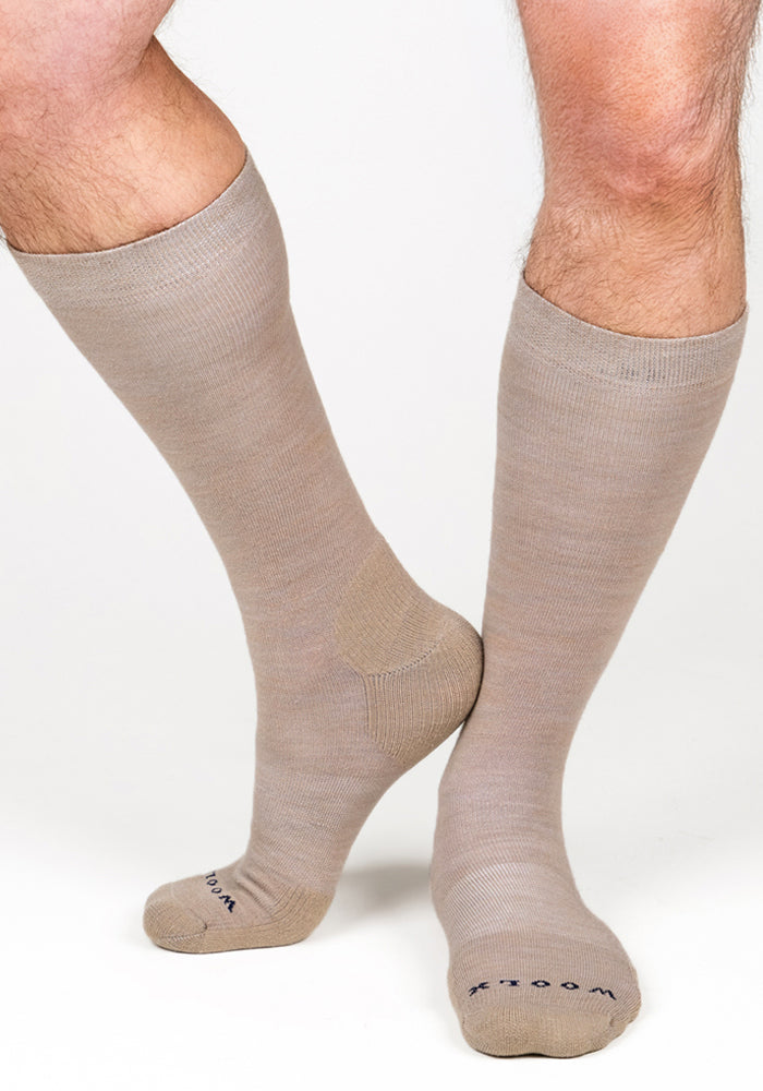 A person wears Woolx Mens Chandler Mid Calf Socks in Simply Taupe, which reach just below the knees with toes pointed slightly inward. The background is plain white.