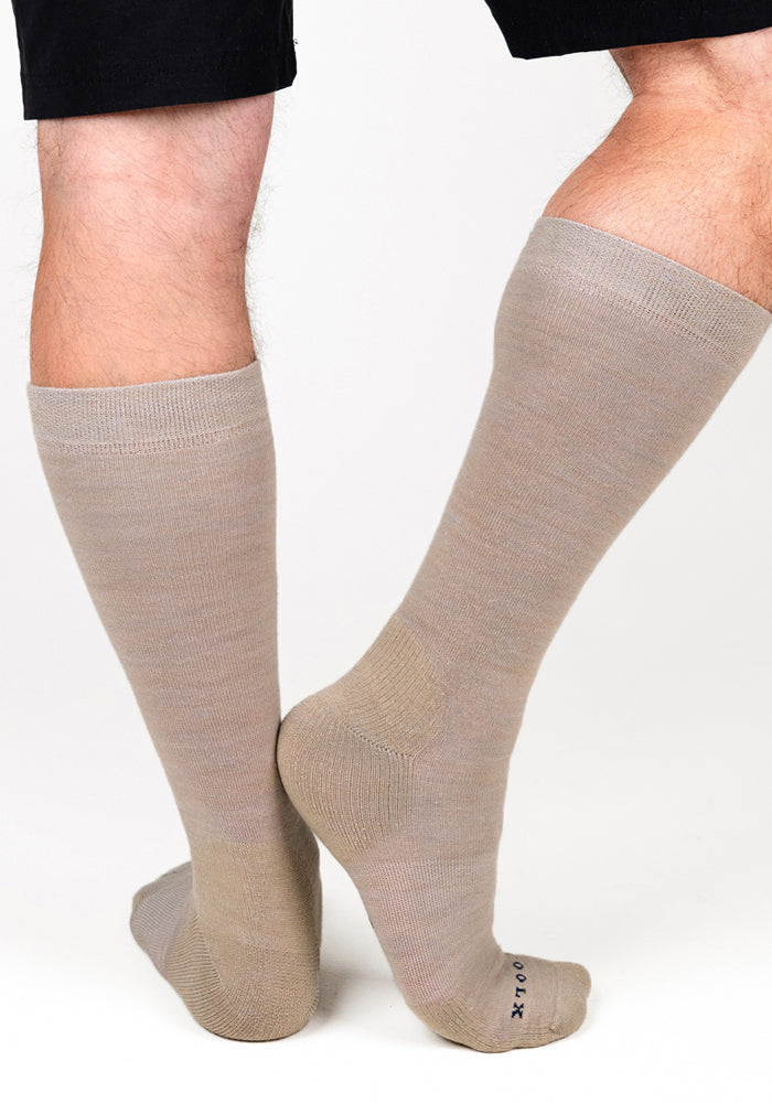 A person models Woolx Mens Chandler Mid Calf Socks in Simply Taupe, standing with one foot flat and the other pointed to display the side and back of the socks against a white background.