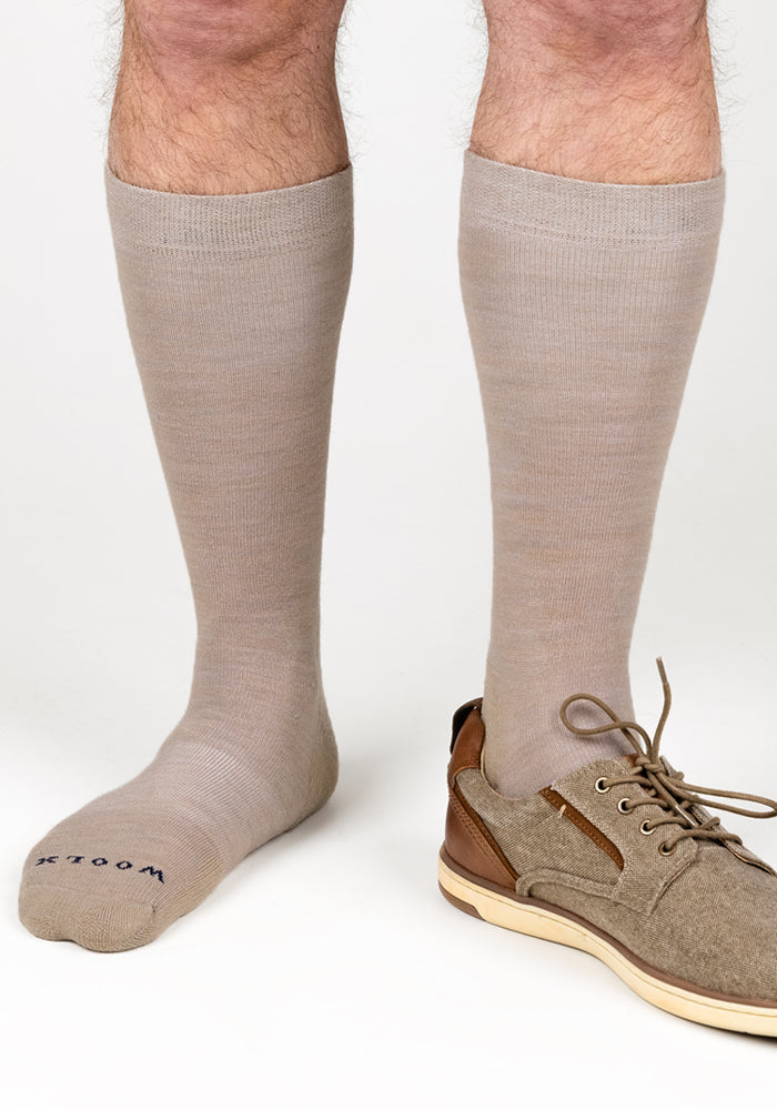 A person wearing Woolx Mens Chandler Mid Calf Socks in Simply Taupe, with one foot in a beige and brown lace-up shoe and the other bare, stands on a white background.