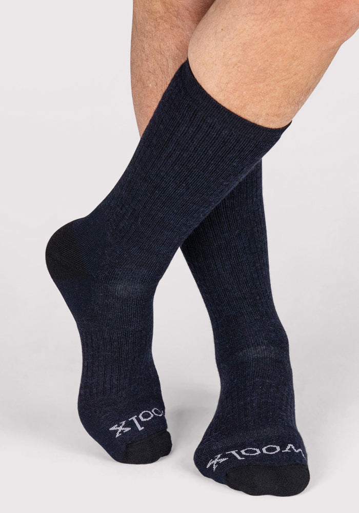 A pair of legs wears Woolx Mens Core Crew Sock - Navy, featuring black heels and toes, with "1004" and a small white logo on the toes. The background is plain and light-colored.
