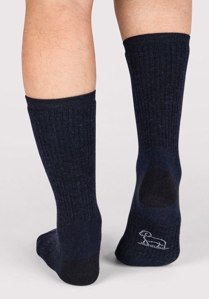 A person wears Woolx Mens Core Crew Sock - Navy, standing on a light surface. The crew sock, visible from mid-calf down, features a small white outline of a dachshund dog on the heel.