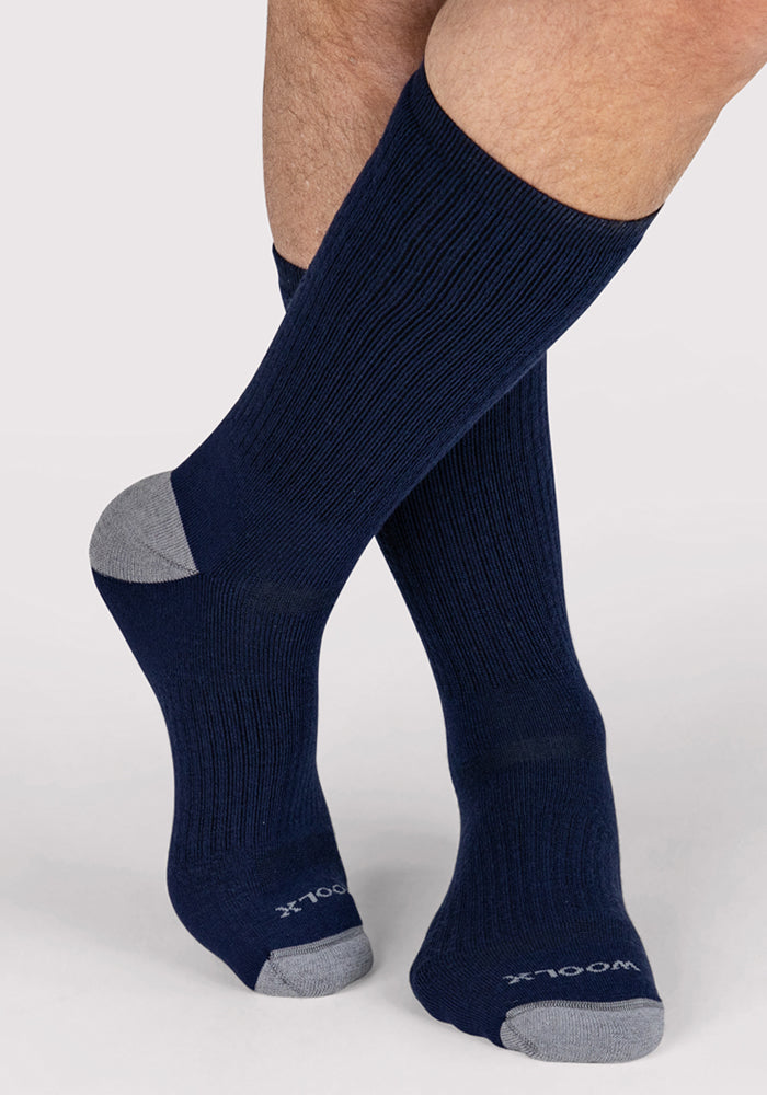 A person wearing Woolx Mens Core Crew Socks in Deep Navy with gray heels and toes, "100X" printed in white on the toe, stands on a light background with one leg crossed over the other.