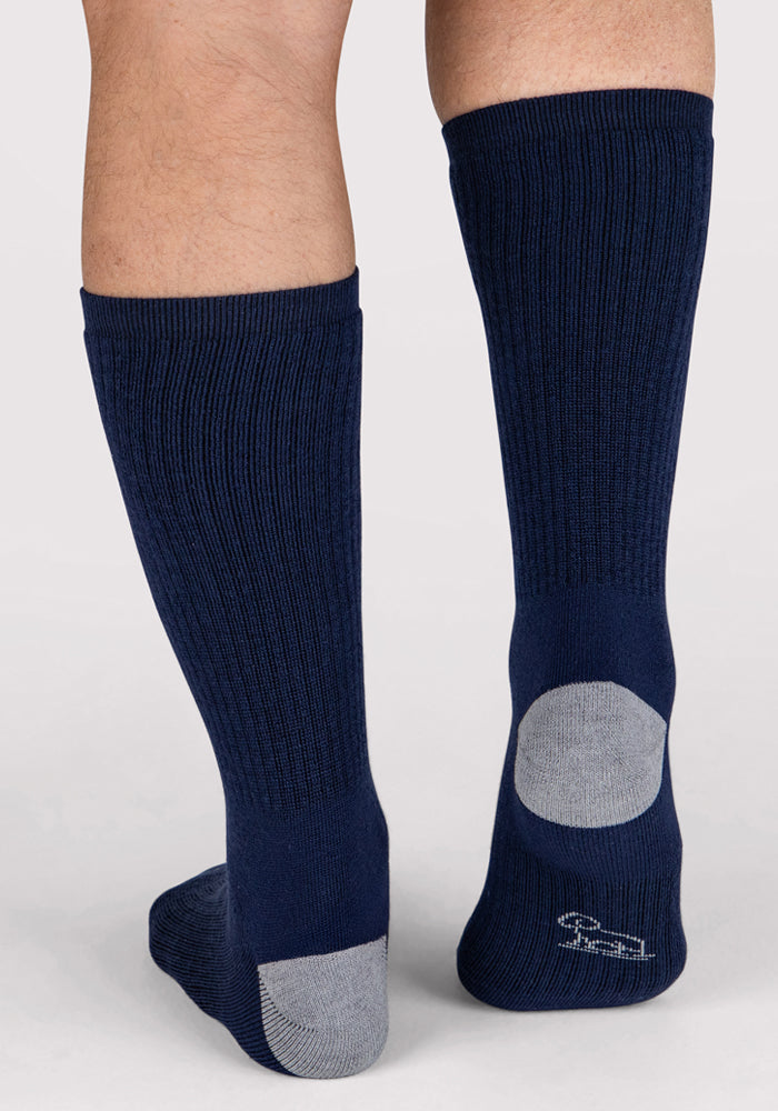 A pair of legs wearing Woolx Mens Core Crew Socks in Deep Navy, shown from the back. These mid-calf socks have cushioned soles, gray heels and toes, and a small white embroidered logo near the heel for a stylish touch.