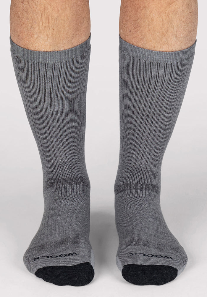 A pair of feet wearing Woolx Mens Core Crew Socks in grey with black toes, standing on a light surface. These ribbed, moisture-wicking crew socks reach the lower calves and feature text on the toes.