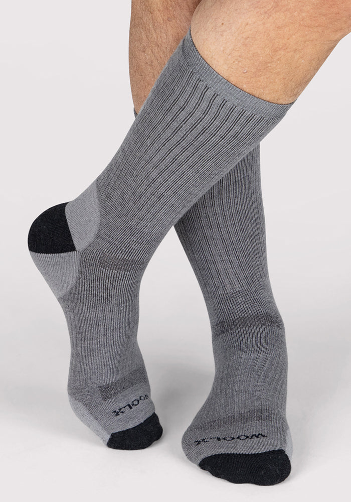 A person stands on a light background wearing Woolx Mens Core Crew Socks in grey, featuring black toe and heel accents, moisture-wicking fabric, arch support, and text on the toes.