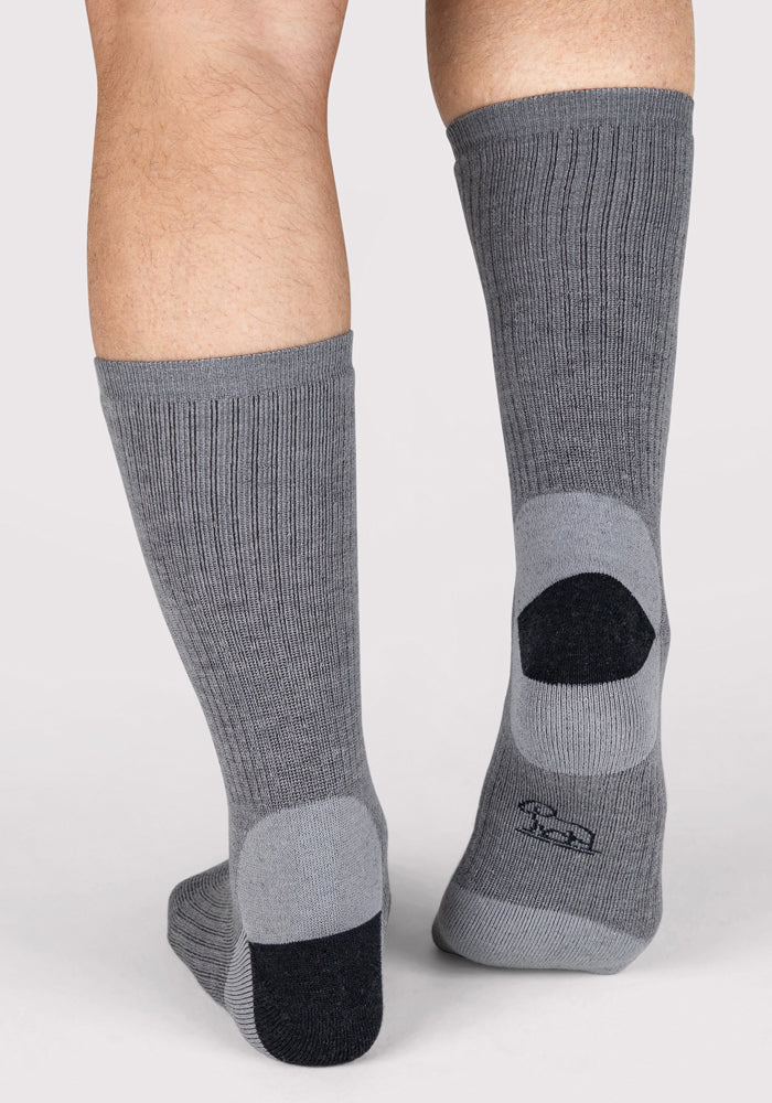 A person wears Woolx Mens Core Crew Socks - Grey, featuring arch support and ribbed merino wool with dark gray heels and black toe/heel caps, standing on a light background shown from the back calves down.