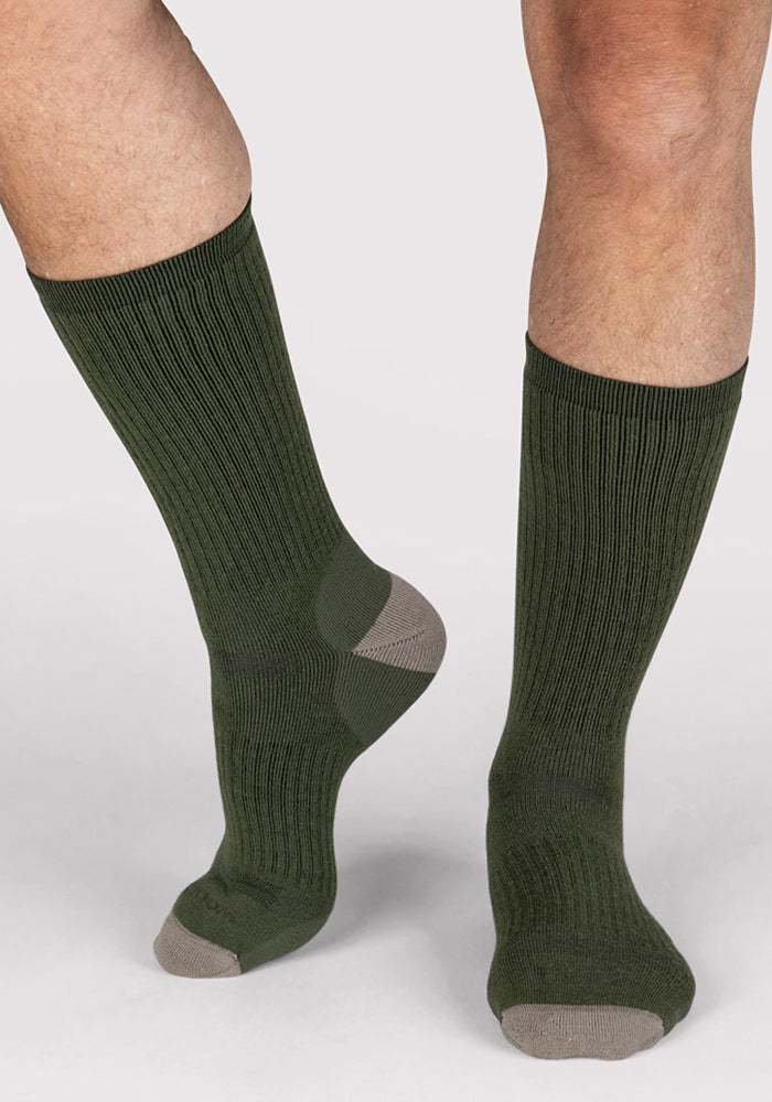 A person stands on a light background wearing Woolx Mens Core Crew Socks in Winter Moss. The ribbed, dark green merino wool socks feature light gray toes and heels. Only the lower legs and feet are visible.