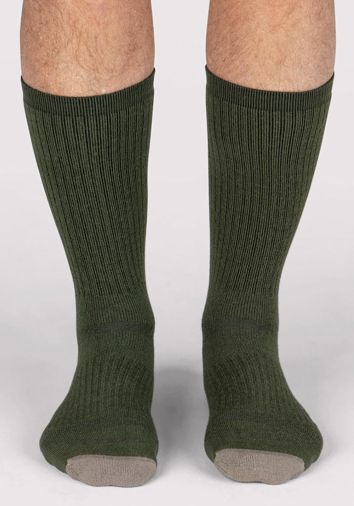 A pair of legs wearing Woolx Mens Core Crew Socks in Winter Moss with ribbed dark green merino wool and light gray toes, standing on a plain light background.