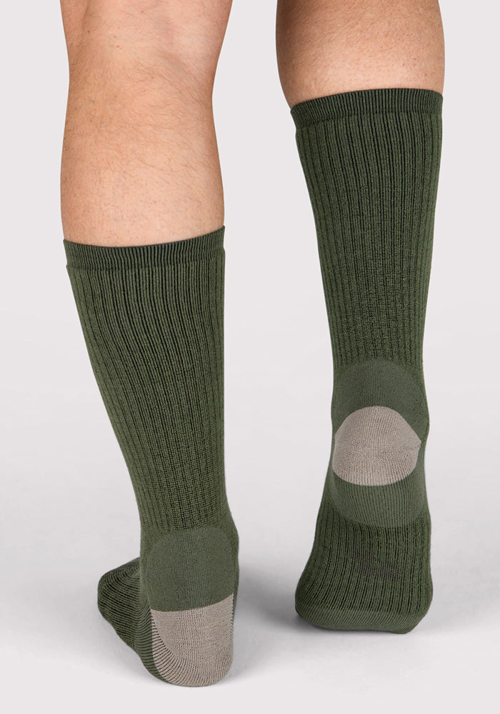 A close-up of two legs wearing Woolx Mens Core Crew Socks in Winter Moss, featuring green ribbed merino wool with gray heel patches, standing on a light background.