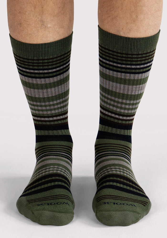 A pair of feet wears Woolx Mens Everyday Crew Socks in Winter Moss Stripe—green, black, and gray Merino wool crew socks with geometric horizontal stripes. These cushioned, moisture-wicking socks reach mid-calf against a light background.