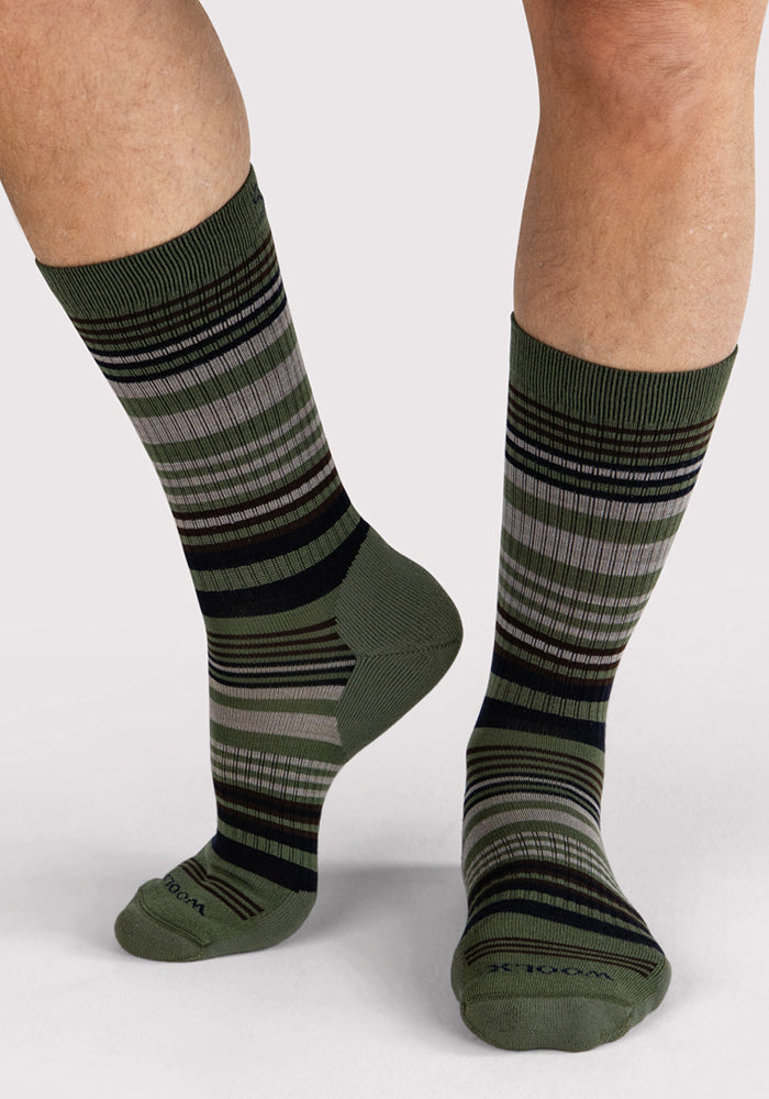 A person wears Woolx Mens Everyday Crew Socks in Winter Moss Stripe—moisture-wicking green socks featuring black, gray, and dark green stripes—while standing on a light surface. Only their lower legs and feet are shown.