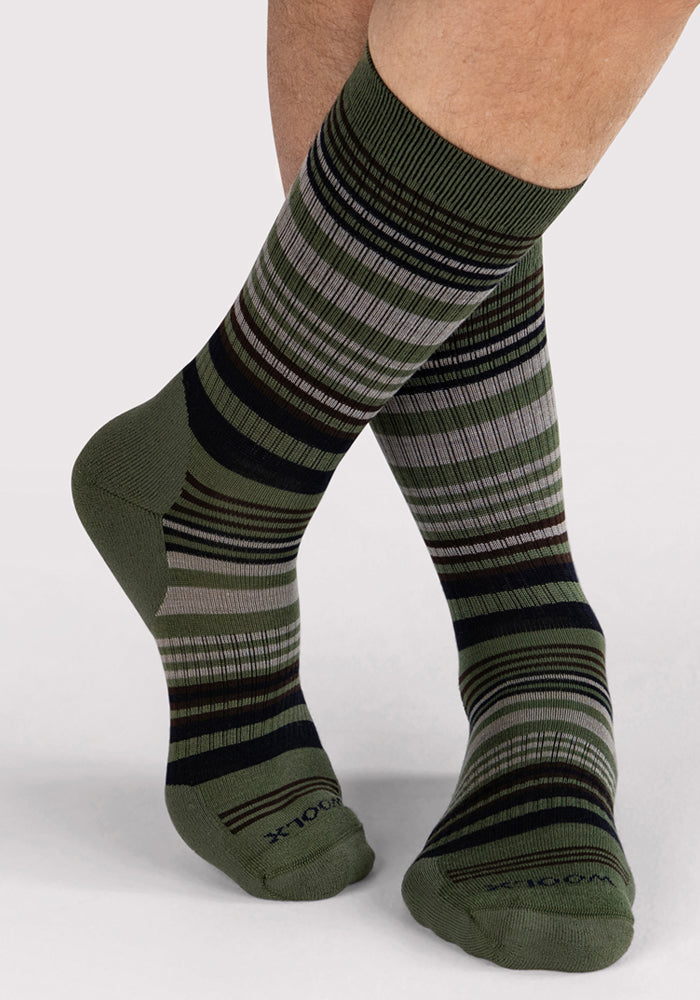 A person wearing Woolx Mens Everyday Crew Socks in Winter Moss Stripe—moisture-wicking green socks with black, brown, and gray horizontal stripes—stands with legs crossed on a light background.