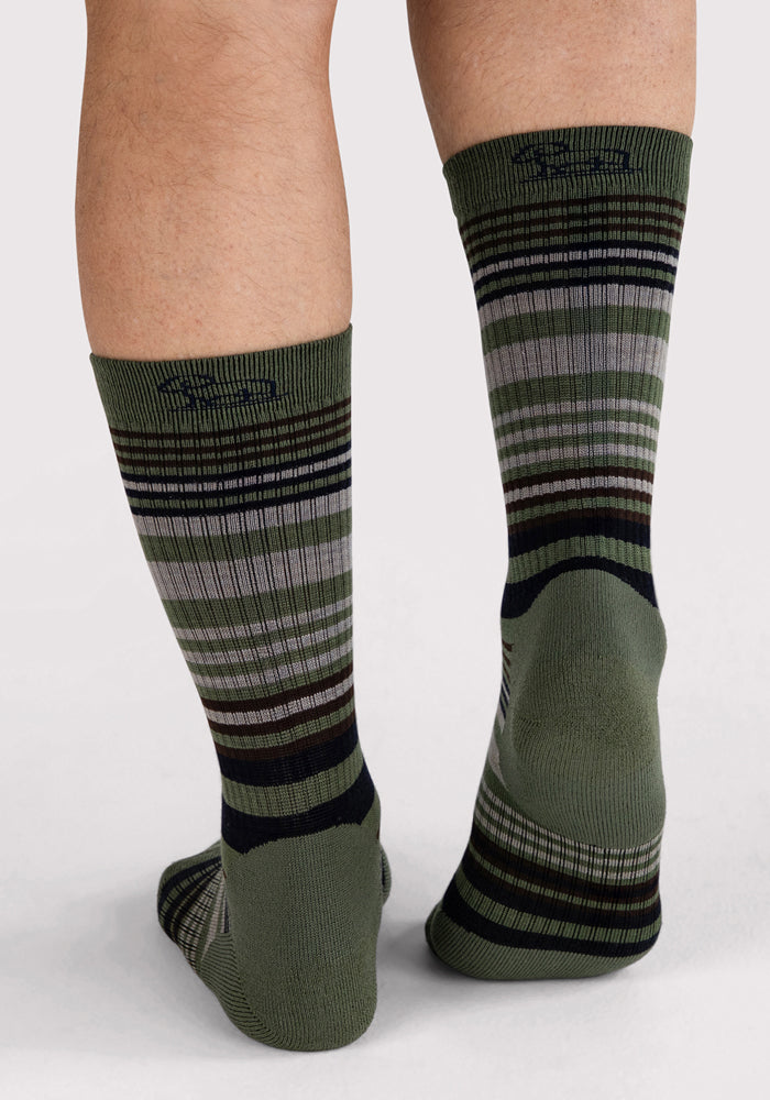 A person stands on a white surface, shown from behind, wearing Woolx Mens Everyday Crew Socks in Winter Moss Stripe—green moisture-wicking socks featuring black, gray, and white horizontal stripes. Only the lower legs and feet are visible.