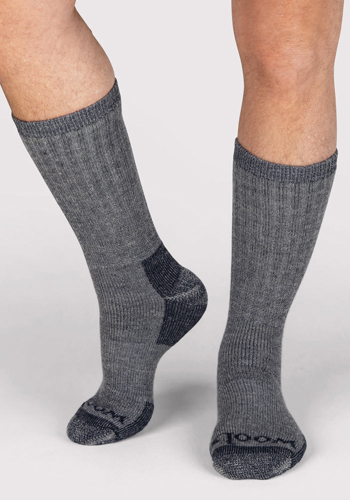 A close-up shows only the lower legs and feet of a person wearing Woolx Mens Extreme Crew Socks in Charcoal Heather, featuring ribbed mid-calf design with darker heels and toes, standing on a light-colored surface.